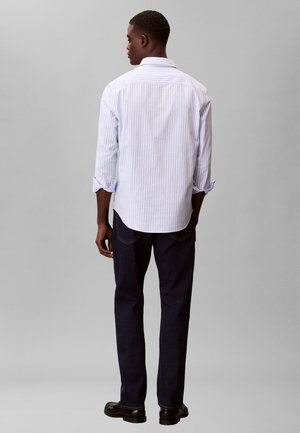 Mies seisoo selin, pukeutuneena vaaleansiniseen raidalliseen nappipaitaan, tummiin farkkuihin ja mustiin kenkiin yksiväristä harmaata taustaa vasten.