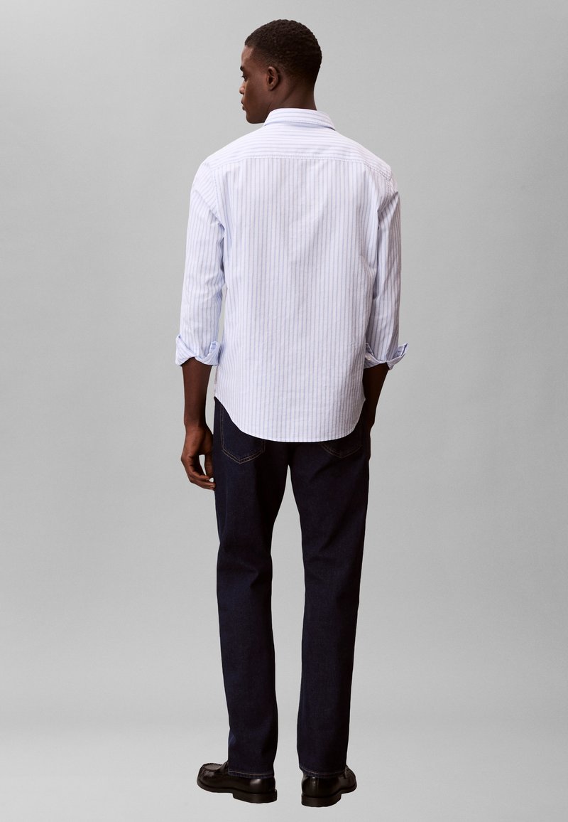 Άνδρας που στέκεται με την πλάτη στραμμένη φορώντας ανοιχτό μπλε ριγέ πουκάμισο με κουμπιά, σκούρο τζιν και μαύρα παπούτσια σε απλό γκρι φόντο.