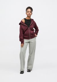 Sweat à capuche zippé bordeaux avec capuche, coupe décontractée et découpe latérale. Associé à un pantalon large gris clair et des chaussures noires. Texture de tissu lisse.