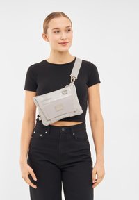 Borsa a tracolla grigia realizzata in materiale lucido, con una tasca frontale dotata di un'etichetta con logo, tracolla regolabile e un design a pannelli distintivo.