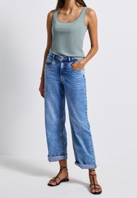 Vaaleansiniset farkut, joissa on käärityt lahkeet, yhdistettynä napakkaan vaaleanvihreään topiin. Malli seisoo yksi käsi taskussa.