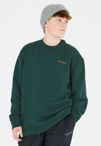 Mörkgrön sweatshirt med ribbad rund hals, broderad "LES'ARCS"-logotyp i orange, bärs med svarta byxor. Tillbehör inkluderar en ljusgrå stickad mössa.