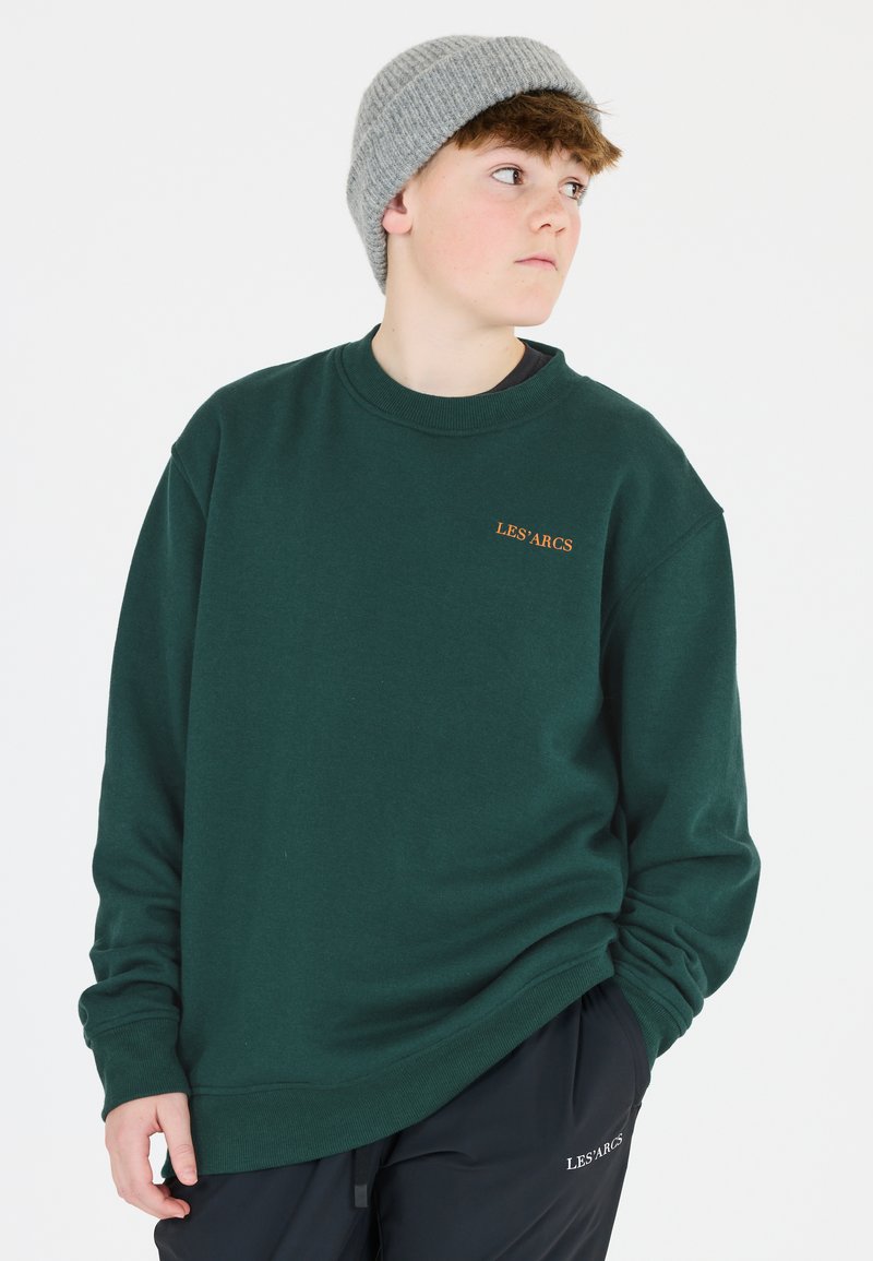Mörkgrön sweatshirt med ribbad rund hals, broderad "LES'ARCS"-logotyp i orange, bärs med svarta byxor. Tillbehör inkluderar en ljusgrå stickad mössa.