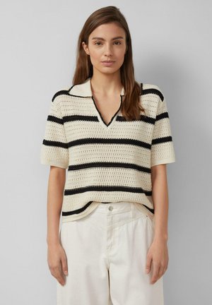 Junge Frau mit glatten braunen Haaren, die ein cremefarbenes und schwarz gestreiftes Strickoberteil mit kurzen Ärmeln und hoch taillierte weiße Hose trägt, steht vor einem grauen Hintergrund.