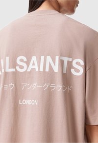 Helles rosa Baumwoll-T-Shirt mit kurzen Ärmeln, auf dem Rücken mit fettem weißen Text, einschließlich "ALLSAINTS" und "LONDON."
