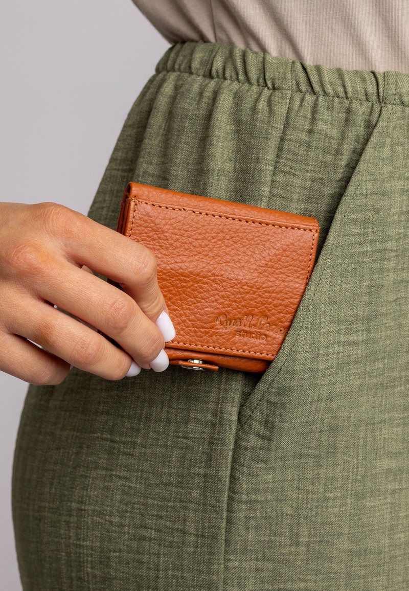 Bruine leren kaarthouder vastgehouden door een hand, deels ingebracht in een groene stoffen pocket. Geweven afwerking met een drukknoopsluiting.