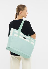 Kvinna med ljusbrunt hår i hästsvans, klädd i svart skjorta och krämfärgade byxor, som bär en stor mintgrön och vitrandig axelväska.