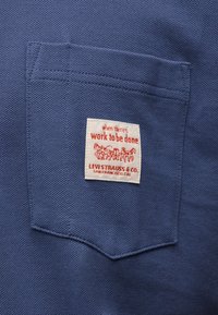 Μπλε υφασμάτινη τσέπη με λευκή ετικέτα που φέρει κόκκινο κείμενο «όταν υπάρχει δουλειά να γίνει» και το λογότυπο της Levi Strauss & Co. με άμαξα που τραβιέται από άλογα.