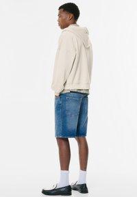 Beige Kapuzenpullover mit lockerem Schnitt, Jeansshorts, weißen Socken und schwarzen Slippern; das Model steht seitlich vor einem schlichten Hintergrund.