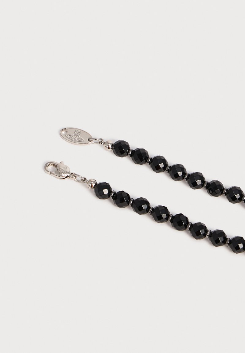 Chaîne de perles facettées noires avec fermoir mousqueton argenté et étiquette métallique ovale avec logo gravé sur fond uni.