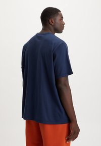 Tricou din bumbac albastru marin, cu croială lejeră, mâneci scurte și decolteu rotund, prezentat din spate, asortat cu pantaloni portocalii.