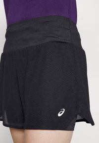 Primer plano de una persona que lleva pantalones cortos deportivos de malla negra con un logo blanco de Asics en el lado inferior derecho y una camiseta morada metida por dentro.