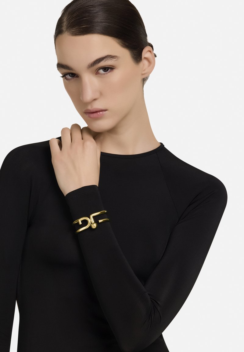 Kuldne käevõru, millel on sfääriline aksent ja voolujooneline disain, kantud modellil, kellel on seljas must pika varrukaga top. Sile tekstuur, minimalistlik stiil.