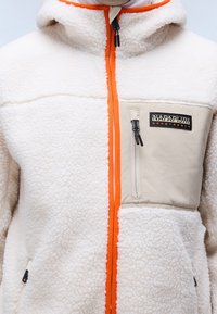 Vitt sherpa-fleecetröja med orange dragkedja, beige bröstficka med vertikal dragkedja och Napapijri Geographic-logotyp.