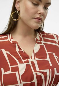 Blusa a maniche corte con scollo a V, a motivo rosso e crema. Presenta forme geometriche astratte e una texture di tessuto morbida. Orecchini dorati con un accento di perla.
