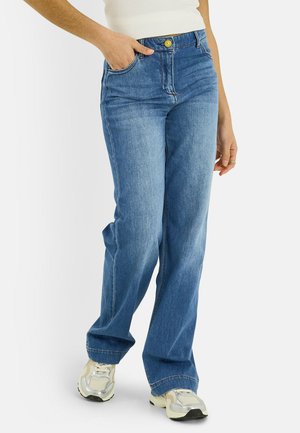 Personne portant un jean droit bleu, des baskets blanches, et un haut blanc, une main dans la poche et l'autre détendue sur le côté.