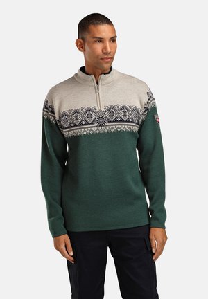 Mann iført grønn og beige genser med nordisk mønster og svarte bukser, står med hendene avslappet langs siden, ser mot høyre.