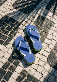 Μπλε σαγιονάρες Havaianas με λευκές λουρίδες πάνω σε μαύρο και άσπρο δαπέδο από ψηφιδωτό με σκιές που πέφτουν στην επιφάνεια.