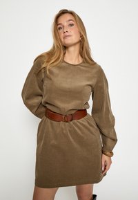 Braunes Cordkleid mit langen Ärmeln, rundem Ausschnitt und tailliertem Schnitt. Ein brauter Ledergürtel mit runder Schnalle betont die Silhouette.