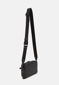 Svart crossbody-veske laget av teksturert materiale, med glidelåslukking, justerbar stropp og en liten tag-detalj. Kompakt rektangulær form.