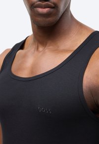 Κοντινό πλάνο ενός ατόμου που φοράει μαύρο ριμπ τοπ χωρίς μανίκια με την επιγραφή "BOSS" κεντημένη στο στήθος, παρουσιάζοντας λεπτομέρειες του λαιμού και των ώμων.