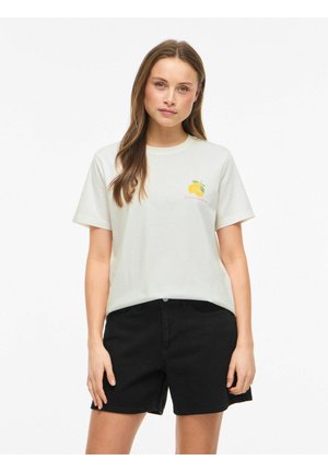 Kvinde iført en hvid t-shirt med et citronmotiv og tekst, kombineret med sorte denimshorts, stående mod en ensfarvet hvid baggrund.