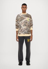 Sweatshirt med zebramönster i beige och svart, med en gul logotyp. Matchad med mörkgrå byxor, accentuerad med en färgglad grafik.