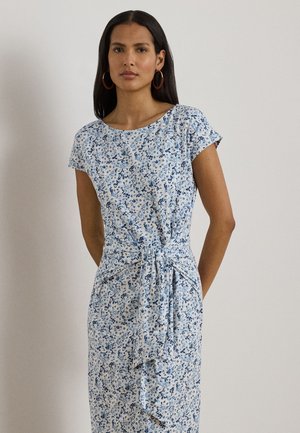 Kobieta o długich ciemnych włosach, nosząca kolczyki w kształcie koła oraz białą sukienkę z krótkim rękawem, niebieskim kwiatowym wzorem i wiązanym paskiem w talii.