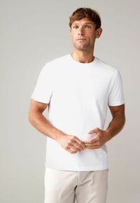 Weißes T-Shirt mit kurzen Ärmeln und Rundhalsauschnitt, aus glattem Stoff. Helle Hose sichtbar. Model steht mit den Händen in der Taille.