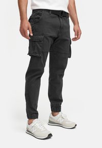 Homme portant un pantalon cargo noir avec des poches latérales, des baskets blanches et une chemise blanche, debout devant un fond blanc uni.