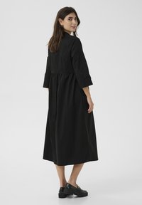 Abito midi nero con maniche a tre quarti, corpetto aderente e vita arricciata. Textura liscia e design minimalista. Abbinato a scarpe nere chunky.