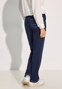 Jeans in denim blu scuro con cuciture bianche, caratterizzati da un taglio dritto e tasche posteriori. Abbinati a sneaker bianche su uno sfondo chiaro.
