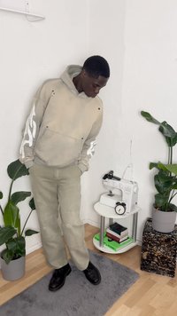 Moletom oversized bege com gráficos de chamas brancas nas mangas, combinado com calças de tom claro e sapatos pretos. Ambiente interior com plantas.