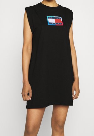 Henkilö, jolla on musta hihaton mekko, jossa on rinnan kohdalla "Tommy Jeans" -logoplässi punaisena, valkoisena ja sinisenä, seisoo yksiväristä valkoista taustaa vasten.
