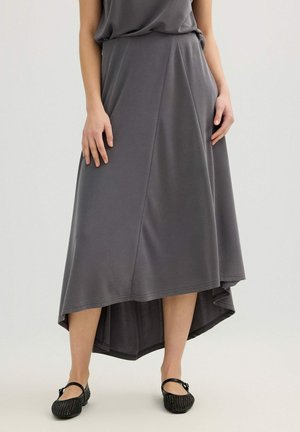 Mujer con falda midi asimétrica gris y bailarinas negras Mary Jane, de pie con las manos relajadas a los lados.