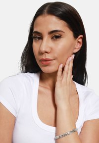 Bracciale a catena in argento al polso, caratterizzato da ampi anelli con una finitura lucida. Indossato con un top bianco aderente, con il tono della pelle visibile.