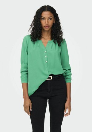 Grüne Bluse mit Knöpfen, tiefem V-Ausschnitt, langen Ärmeln und lockerer Passform. Strukturierten Stoff, kombiniert mit hochtaillierten schwarzen Jeans und einem Gürtel.