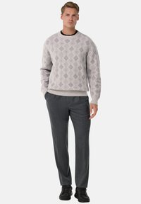 Man som står, iklädd en ljusgrå mönstrad tröja, mörkgrå byxor och svarta sneakers, poserar mot en enkel vit bakgrund.