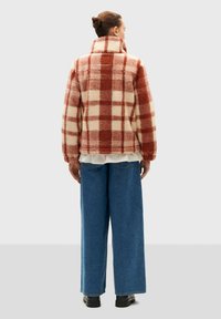 Red and cream plaid fleece jacket with a high collar, wide sleeves, and textured fabric, paired with loose denim trousers.