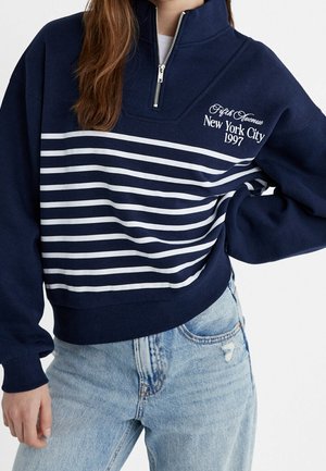 Kobieta ubrana w granatową bluzę z zamkiem do połowy, z białymi poziomymi paskami i napisem "Fifth Avenue New York City 1997", w połączeniu z jasnoniebieskimi dżinsami.