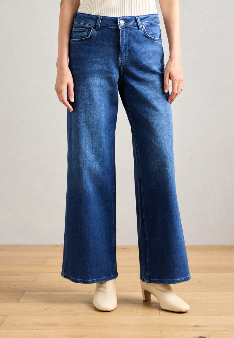 Weite blaue Jeans aus Denim mit Knopf- und Reißverschluss, frontalen Taschen und glatter Textur. Kombiniert mit beigen Schuhen mit Absatz.