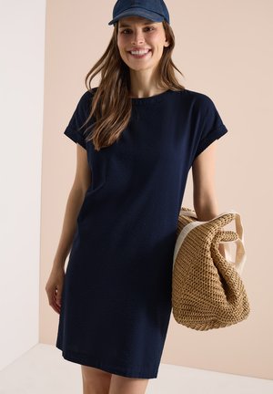 Lächelnde Frau trägt ein marineblaues Kleid und eine Jeanskappe, hält eine große geflochtene Strohtasche und steht vor einem beigen und weißen Hintergrund.