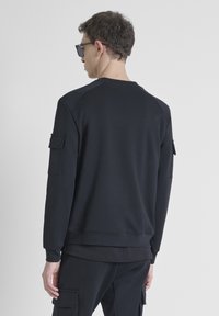Felpa nera con una texture liscia, presenta maniche raglan e due tasche utili sulle braccia, indossata con pantaloni neri abbinati.