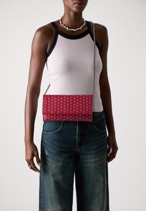 Pochette en simili cuir rouge avec des clous argentés, au design rectangulaire, surface texturée et bandoulière en chaîne. Portée avec un jean large bleu marine.