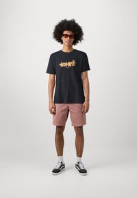 Black cotton t-shirt with orange floral design, paired with pink cotton shorts and black-and-white sneakers. Model with curly hair and sunglasses.
