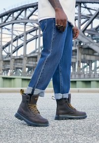 Botas de cuero marrón hasta el tobillo con cordones amarillos, suela robusta y una pestaña de agarre. Llevadas con jeans azules arremangados bajo un fondo industrial.