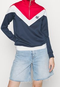 Felpa navy con design a zigzag rosso e bianco, colletto alto, mezza zip e logo. Abbinata a pantaloni corti in denim blu chiaro con orlo sfrangiato.