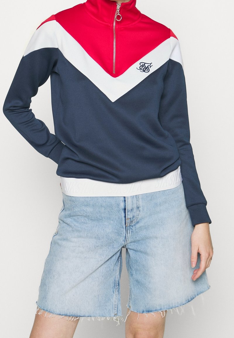 Felpa navy con design a zigzag rosso e bianco, colletto alto, mezza zip e logo. Abbinata a pantaloni corti in denim blu chiaro con orlo sfrangiato.