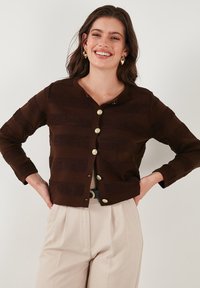 Cardigan marron avec des rayures horizontales texturées, un col rond et de grands boutons dorés. Porté sur un pantalon beige clair.