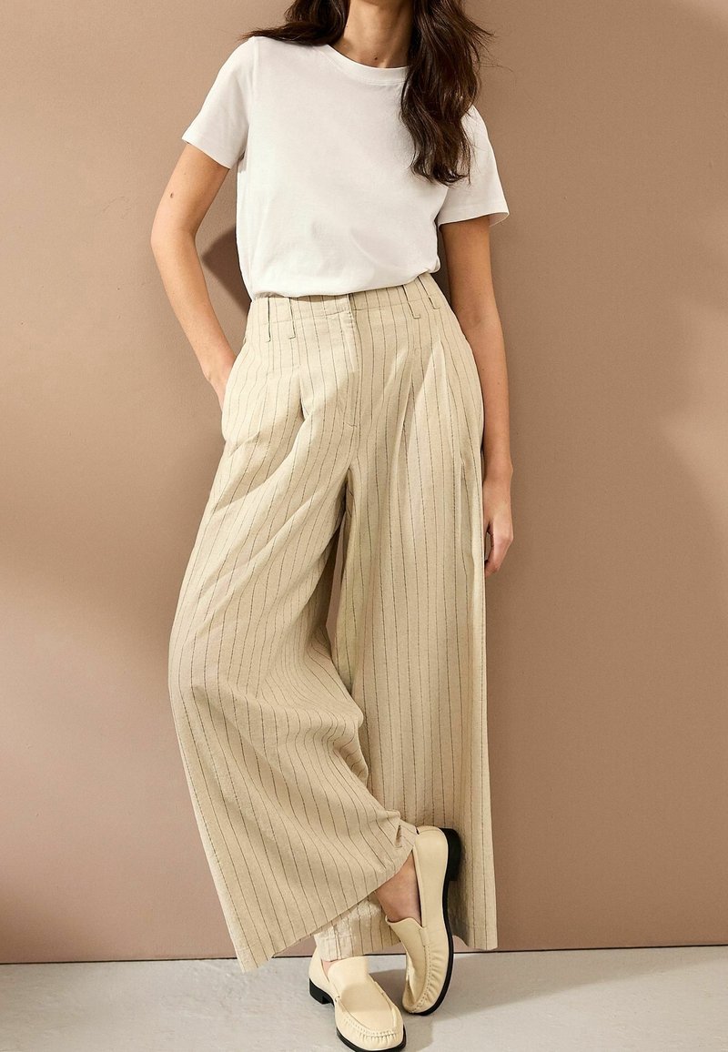 Femme portant une chemise blanche à manches courtes, un pantalon beige à larges jambes à fines rayures et des mocassins beiges, debout contre un mur beige.